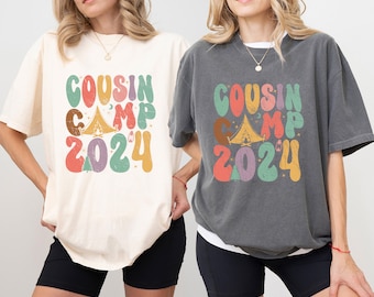 Può includere: Due t-shirt con il testo "COUSIN CAMP 2024" in un font retrò con una grafica di tenda da campeggio e falò. Una maglietta è color crema e l'altra è grigio scuro.