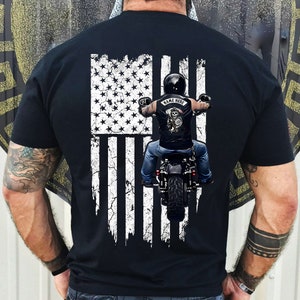 May include: Black t-shirt featuring a distressed American flag design with a biker riding a motorcycle. The flag is white and grey with red stripes. The biker is wearing a helmet and a vest with the text "NAME HERE".