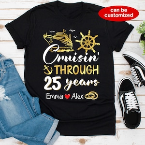 Op de afbeelding: Zwart T-shirt met gouden tekst die "Cruisin' Through 25 years" luidt met een afbeelding van een schip, anker en stuurwiel. De namen "Emma" en "Alex" staan onder de tekst met een rood hart en gouden ringen.