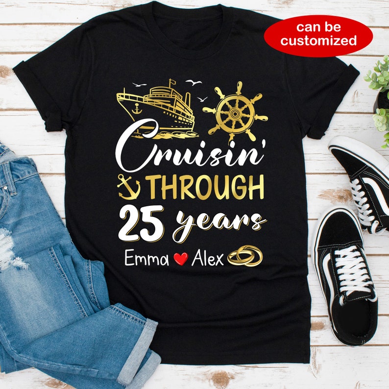 Op de afbeelding: Zwart T-shirt met gouden tekst die "Cruisin' Through 25 years" luidt met een afbeelding van een schip, anker en stuurwiel. De namen "Emma" en "Alex" staan onder de tekst met een rood hart en gouden ringen.