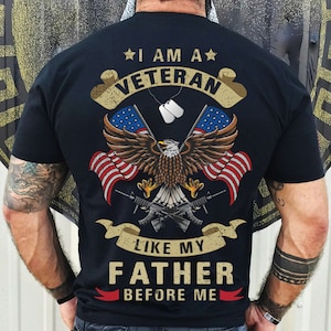 Puede incluir: Camiseta negra con un dise&ntilde;o gr&aacute;fico desgastado que presenta un &aacute;guila con las alas extendidas sosteniendo dos banderas americanas. El texto "I am a Veteran like my Father before me" est&aacute; impreso en la camiseta.