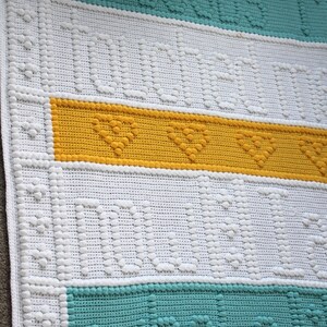 May include: A white crocheted baby blanket with a yellow, teal and white striped pattern. The blanket has a textured design with raised bumps.