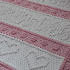 May include: A white and pink crocheted baby blanket with a heart pattern. The blanket is made with soft yarn and has a textured surface.