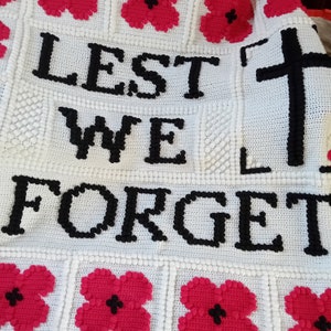 May include: White crochet blanket with the words "Lest We Forget" and a cross in black yarn. The blanket is decorated with red poppy flowers.