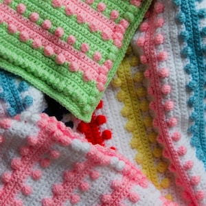 Pode incluir: Close-up de um cobertor de crochê multicolorido com diferentes padrões e texturas. O cobertor é feito com uma variedade de cores, incluindo rosa, verde, azul, amarelo e branco. O cobertor tem uma superfície texturizada com saliências elevadas.