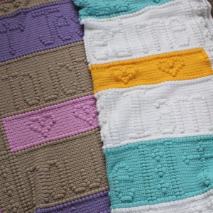 May include: A multi-coloured crocheted blanket with a textured heart pattern. The blanket is made up of stripes of purple, brown, pink, white, yellow, and turquoise.