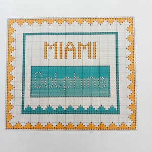 May include: A cross-stitch pattern with a yellow border and a blue and white interior. The pattern reads "Miami Dolphins".