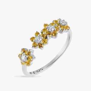 May include: A silver ring with a floral design. The ring features three flower clusters with yellow and white gemstones. The band is open, with a flower on each end. The ring is set against a white background.