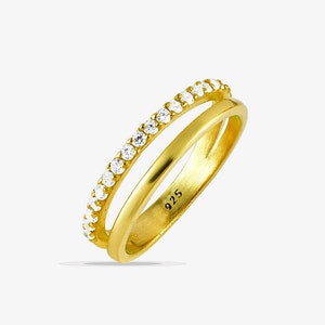 May include: A gold-colored ring with a double-band design. The top band is adorned with small, clear gemstones. The ring is marked with "925". The ring is set against a white background.