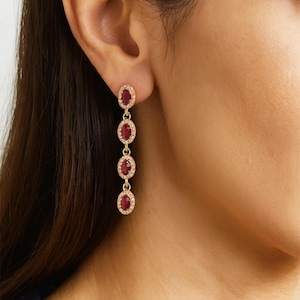 May include: Gold-toned drop earrings featuring oval-shaped, deep red gemstones. Each gemstone is surrounded by a halo of small, clear crystals. The earrings dangle from a small oval at the top, creating a sophisticated, elegant look.