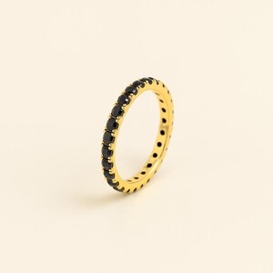 May include: A gold ring featuring a full band of small, black gemstones. The ring is displayed against a pale, off-white backdrop, accentuating the contrast between the gold and black. The ring's design is simple and elegant.
