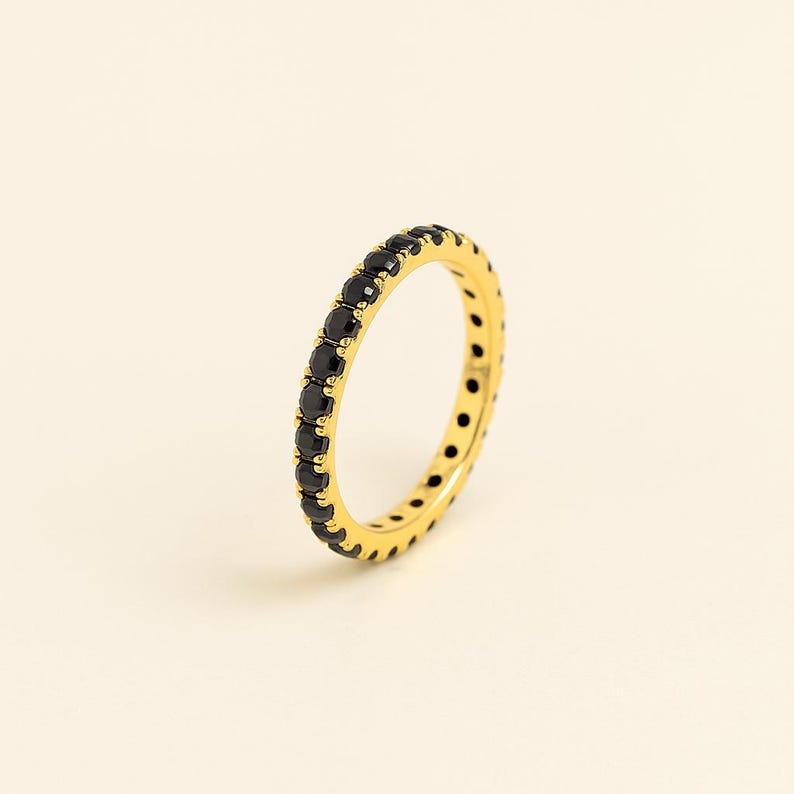 May include: A gold ring featuring a full band of small, black gemstones. The ring is displayed against a pale, off-white backdrop, accentuating the contrast between the gold and black. The ring's design is simple and elegant.