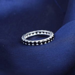 May include: A silver ring featuring a continuous row of small, black gemstones. The ring is displayed on a dark blue fabric, which accentuates the contrast. The gemstones are uniformly set around the ring's circumference.