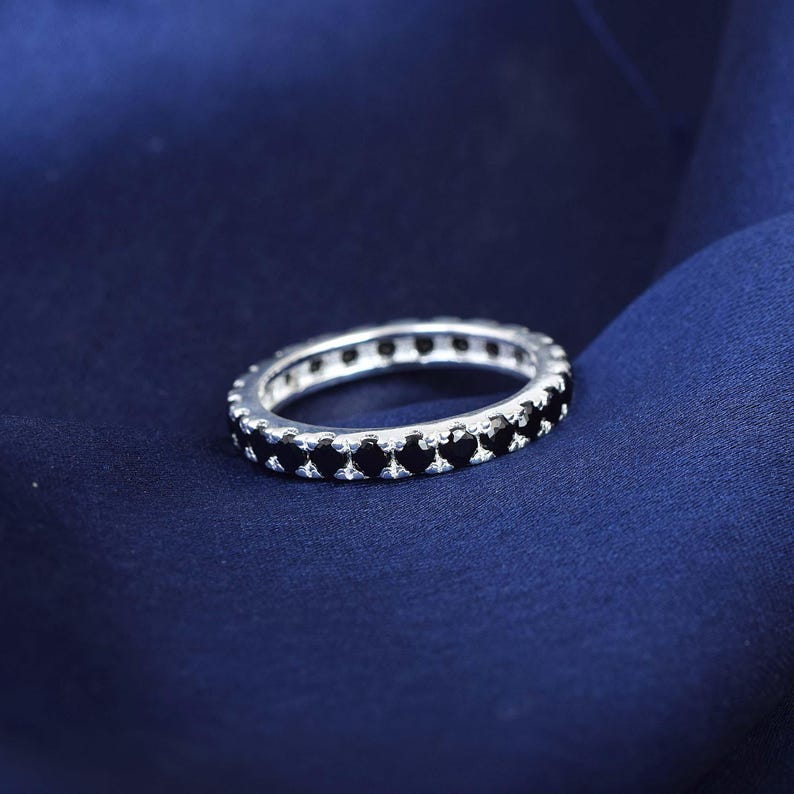 May include: A silver ring featuring a continuous row of small, black gemstones. The ring is displayed on a dark blue fabric, which accentuates the contrast. The gemstones are uniformly set around the ring's circumference.