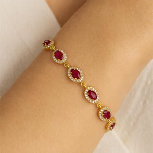 Puede incluir: Pulsera dorada con piedras preciosas rojas oscuras de forma ovalada. Cada piedra preciosa está rodeada de piedras más pequeñas y transparentes. La pulsera está unida por eslabones delicados.