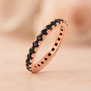 May include: A rose gold ring with a full band of black gemstones. The ring's metal has a warm hue, and the black gemstones are set closely together, forming a continuous line. The background is a soft, blurred gradient of pink and peach tones.