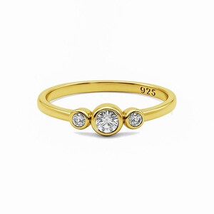 May include: A gold-colored ring with three clear, round gemstones. The ring band is thin and smooth, with the number "925" engraved on the band. The gemstones are set in a bezel setting, with the center stone slightly larger than the side stones.