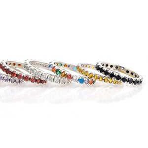 May include: A selection of silver eternity rings, each set with a continuous line of coloured gemstones. The rings display a spectrum of colours, including red, white, green, yellow, blue, and black, offering a vibrant and striking appearance.