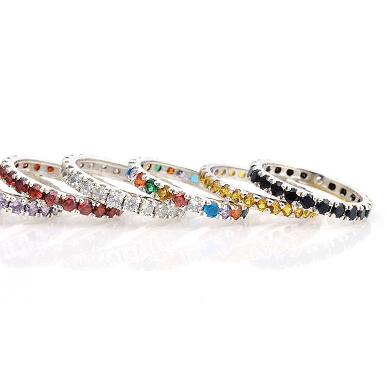 May include: A selection of silver eternity rings, each set with a continuous line of coloured gemstones. The rings display a spectrum of colours, including red, white, green, yellow, blue, and black, offering a vibrant and striking appearance.