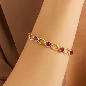 Puede incluir: Un brazalete dorado con eslabones ovalados y circulares, cada uno con una vibrante gema roja. El brazalete se lleva en un brazo, mostrando su diseño y contraste de color. Las gemas son de un rojo intenso.