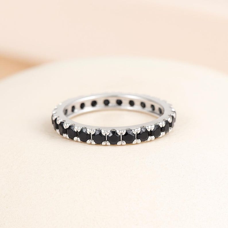 May include: A silver ring featuring a continuous row of small, round, black gemstones. The ring's design is set against a soft, neutral backdrop, accentuating the contrast between the polished metal and the dark stones. It is a full eternity ring.