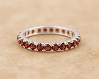 May include: A silver ring with a full band of small, round, deep red gemstones. The gemstones are set closely together, creating a continuous row of colour. The ring is shown on a neutral, textured surface.