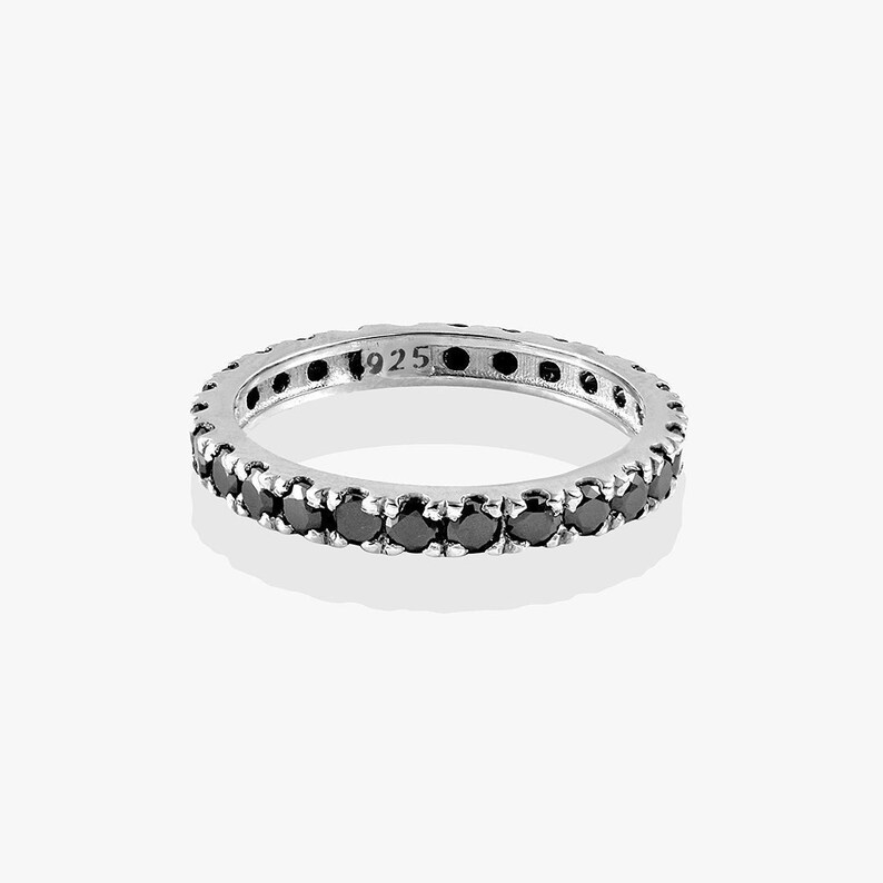 May include: A silver ring featuring a continuous band of small, round black gemstones. The ring is hallmarked with "925" on the inside. The ring is displayed on a white backdrop.