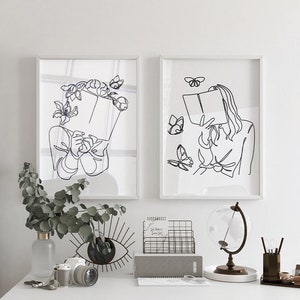 May include: Two framed wall art prints featuring black line art illustrations on white backgrounds. The left print shows a figure holding a book with flowers and a butterfly. The right print depicts a figure reading a book surrounded by butterflies.