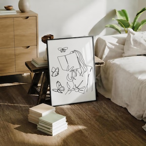 May include: A framed wall art print featuring a black continuous line drawing on a white background. The illustration depicts a person holding an open book in front of their face, surrounded by three butterflies of varying sizes in flight.