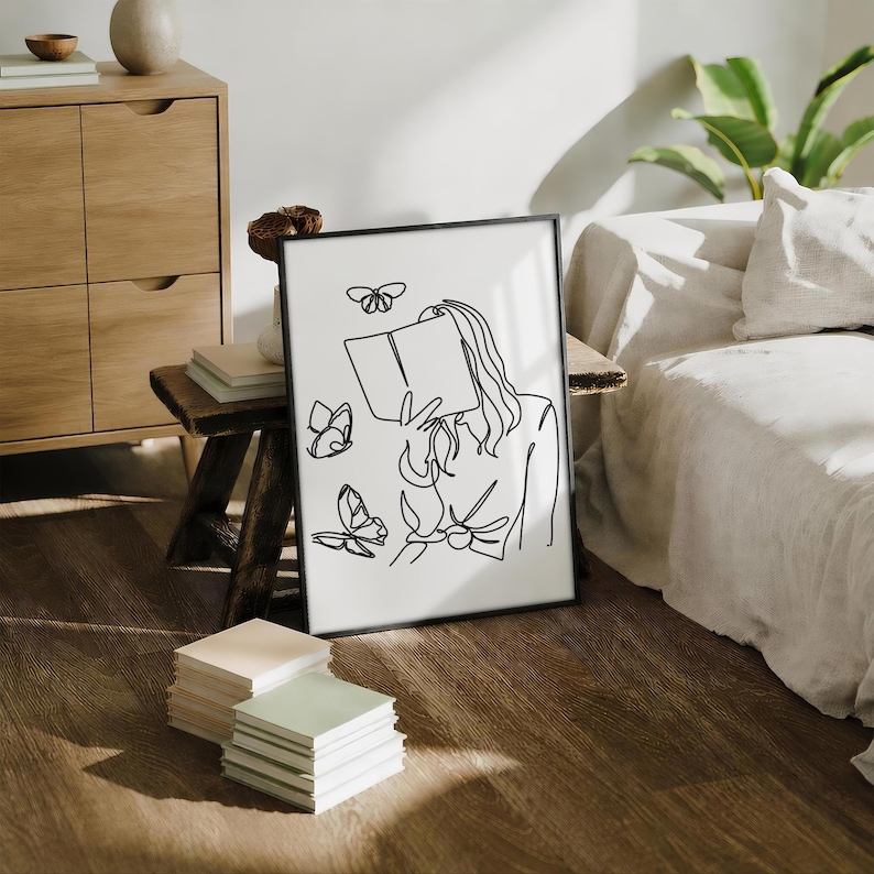 May include: A framed wall art print featuring a black continuous line drawing on a white background. The illustration depicts a person holding an open book in front of their face, surrounded by three butterflies of varying sizes in flight.