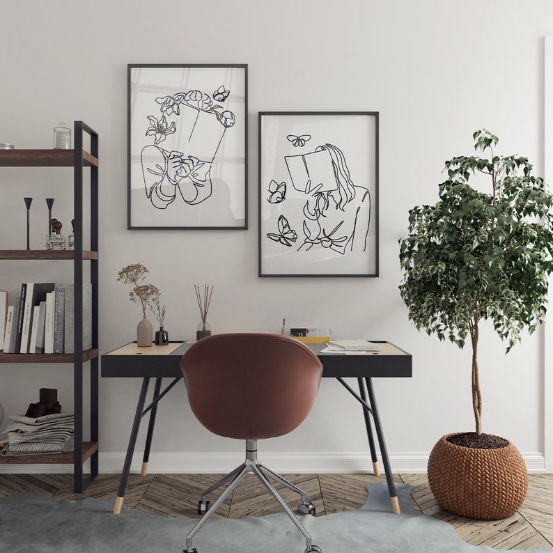 May include: Two framed black and white line art prints hanging on a wall above a desk. The left print shows a person holding a book with flowers, and the right print shows a person reading a book surrounded by butterflies, both in black ink.