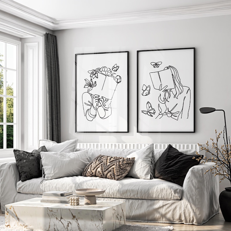 May include: Two framed wall art prints featuring black line art on a white background. The left print shows a figure holding a book with flowers and butterflies around the head. The right print shows a figure holding a book with butterflies.