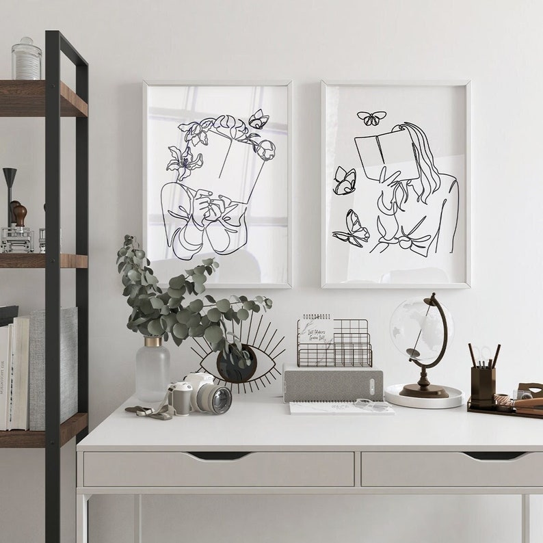May include: Two framed wall art prints featuring black line art illustrations on white backgrounds. The left print shows a figure holding a book with flowers and a butterfly. The right print depicts a figure reading a book surrounded by butterflies.