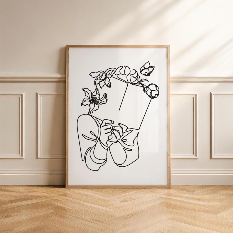 May include: A framed wall art print featuring a black line drawing on a white background. The illustration depicts a person holding an open book in front of their face, with various floral elements blooming from the top of the book.