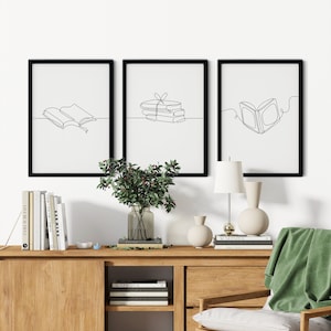 May include: Three black framed prints with white backgrounds and black line drawings of books. The prints are hanging on a white wall above a wooden shelf with books, vases, and a lamp.