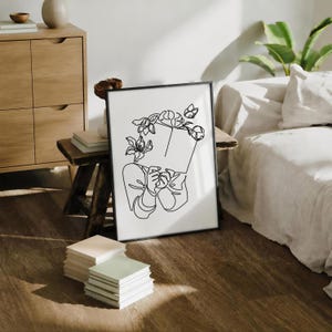 May include: A framed wall art print featuring a black line drawing on a white background. The illustration depicts a person holding an open book in front of their face, with various flowers blooming from the top of the book and around the head area.