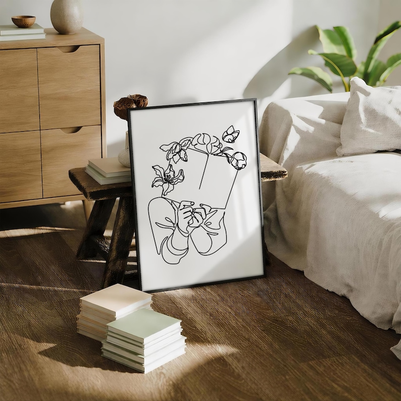 May include: A framed wall art print featuring a black line drawing on a white background. The illustration depicts a person holding an open book in front of their face, with various flowers blooming from the top of the book and around the head area.