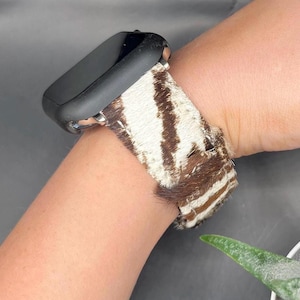 Op de afbeelding: Een zwarte smartwatch met een zebraprint band. De band heeft een patroon van bruine en witte strepen. Het horloge zit om een pols, waarbij de textuur en het ontwerp van de band worden getoond.