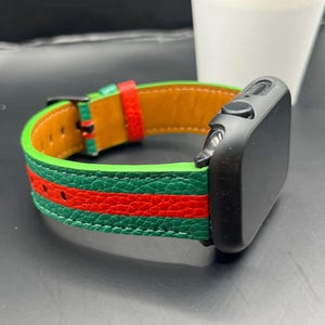 Może przedstawiać: Czarny smartwatch z zielonym, czerwonym i brązowym skórzanym paskiem. Pasek ma zieloną podstawę z czerwonymi paskami i brązowe wnętrze. Zegarek jest prezentowany na ciemnej powierzchni.