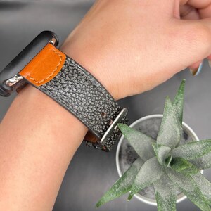 May include: A black and gray textured leather watch band with an orange leather accent is fastened on a wrist. The watch band is secured with a silver buckle. A small succulent plant in a metal pot is in the background.