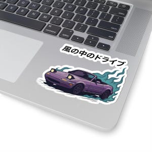 Puede incluir: Una pegatina con un coche descapotable morado con faros emergentes, sobre un fondo de llamas azules estilizadas. El texto japonés "Kaze no naka no doraibu" está encima del coche. La pegatina está en un portátil.