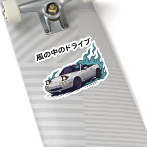 Könnte beinhalten: Ein Aufkleber mit einem weißen Cabrio mit schwarzen Rädern, umgeben von stilisierten blauen Flammen. Der japanische Text "風の中のドライブ" (Kaze no naka no doraibu) befindet sich über dem Auto. Der Aufkleber befindet sich auf einem Skateboard.