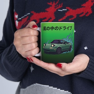 Op de afbeelding: Witte keramische mok met een illustratie van een groene sportwagen en de Japanse tekst "Drive in the Wind" op een groene achtergrond. De auto heeft zijn koplampen aan en stoot blauwe rook uit.