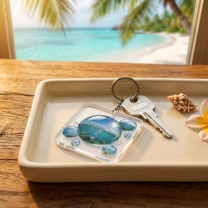 Könnte beinhalten: Ein klarer Acryl-Schlüsselanhänger mit einer Strandszene, einem Schlüssel und einer Muschel auf einem weißen Tablett. Der Schlüsselanhänger hat eine quadratische Form mit einem großen, runden, blauen und transparenten Design in der Mitte, umgeben von kleineren, runden, blauen Designs.