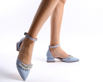 Puede incluir: Sandalias de tacón azul claro con correa en el tobillo y punta afilada. La parte delantera del zapato está adornada con un grupo de pedrería transparente. Los zapatos tienen un tacón bajo y están sobre un fondo blanco.