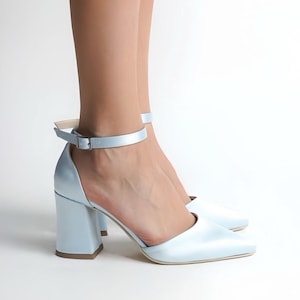 以下が含まれることがあります： アンクルストラップとブロックヒールが付いた、ライトブルーのサテン製ハイヒール。靴はポインテッドトゥで、バックル留めです。ヒールは約7.6cmです。背景は白です。