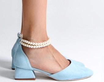 Puede incluir: Zapatos de tacón de ante azul claro con una correa de tobillo de perlas. Los zapatos tienen una punta cuadrada y un tacón de bloque.