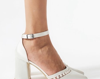 May include: White, closed-toe heels with a block heel and ankle strap. The shoes have a row of small, decorative pearls along the vamp. The heel is approximately 3 inches high.