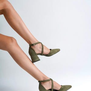 May include: A pair of olive green suede heeled shoes with ankle straps. The shoes have a block heel and a pointed toe.