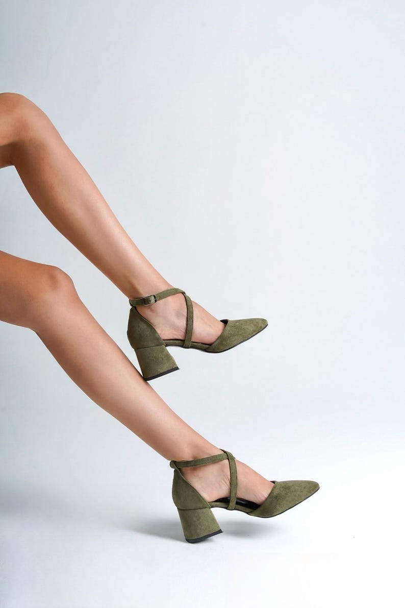 May include: A pair of olive green suede heeled shoes with ankle straps. The shoes have a block heel and a pointed toe.
